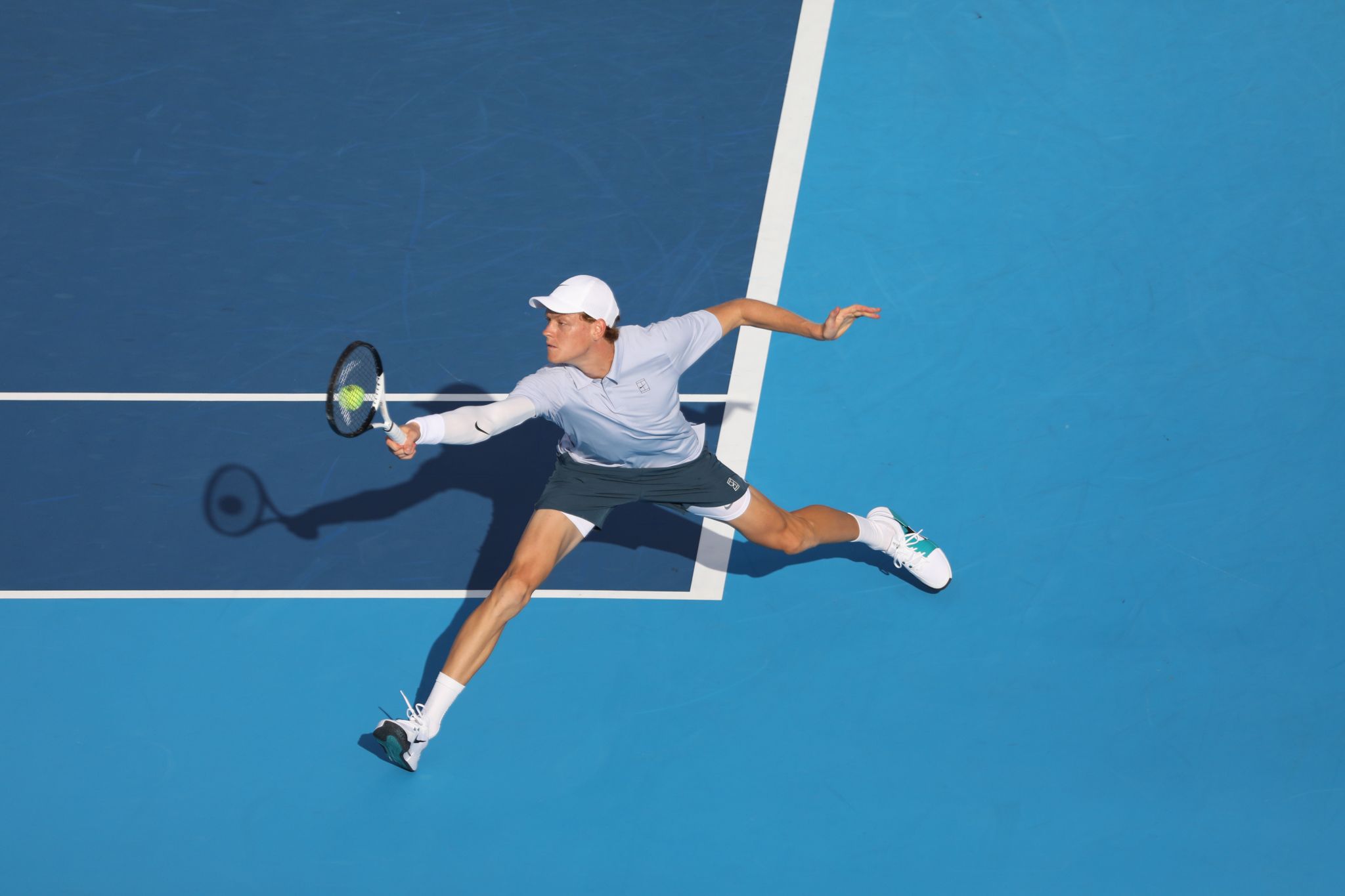 A tennis player in white attire stretches low to return a shot on a blue court during an intense rally.