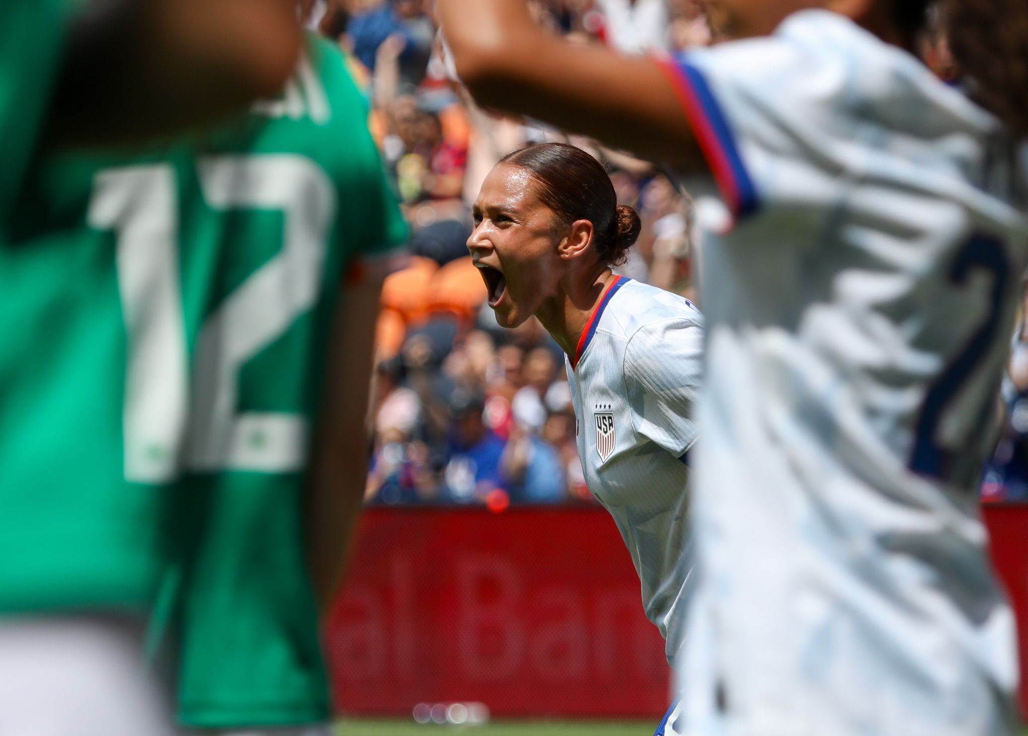 A player in a white jersey celebrates passionately during a match with players in green jerseys visible in the foreground.