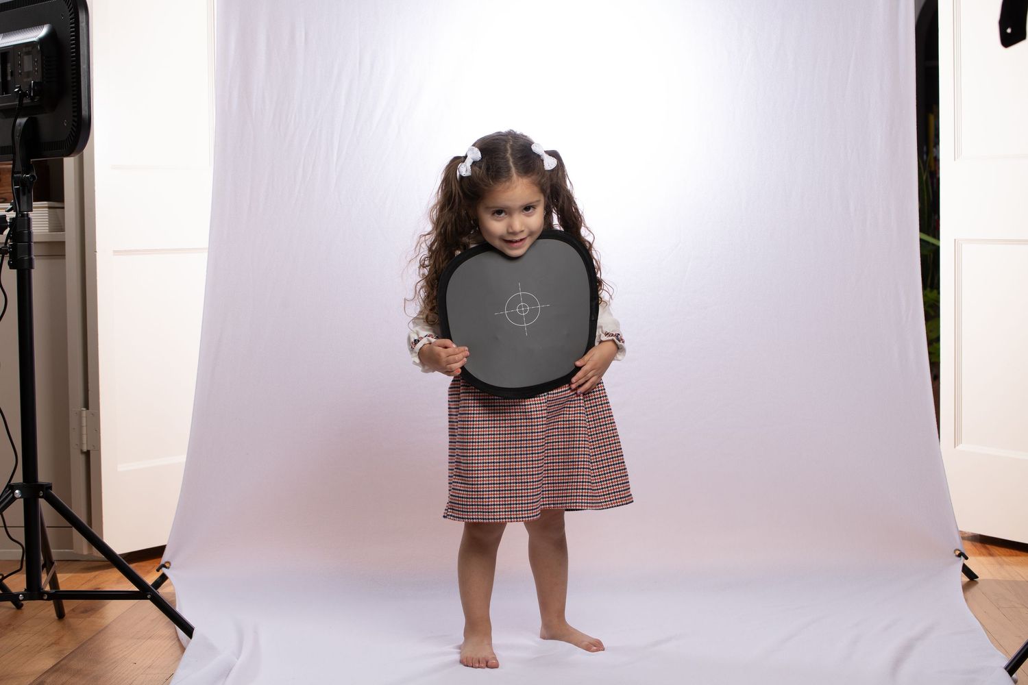 A small child in a dress poses with a circular black reflector in a photo studio setting.