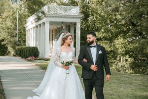 A wedding couple strolls along a garden path with white columns and greenery in the background.