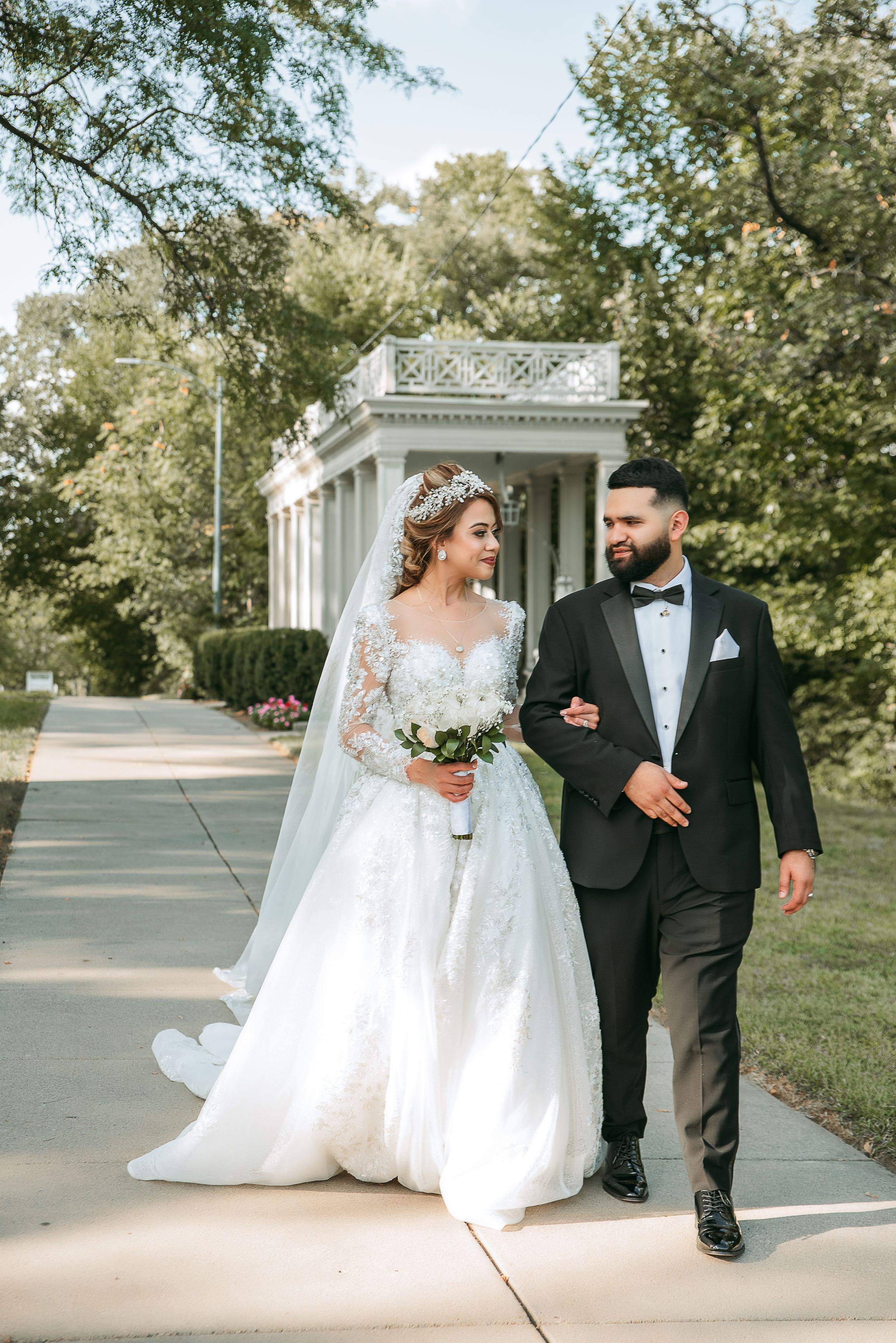 A wedding couple walks together on a scenic outdoor path surrounded by lush trees.