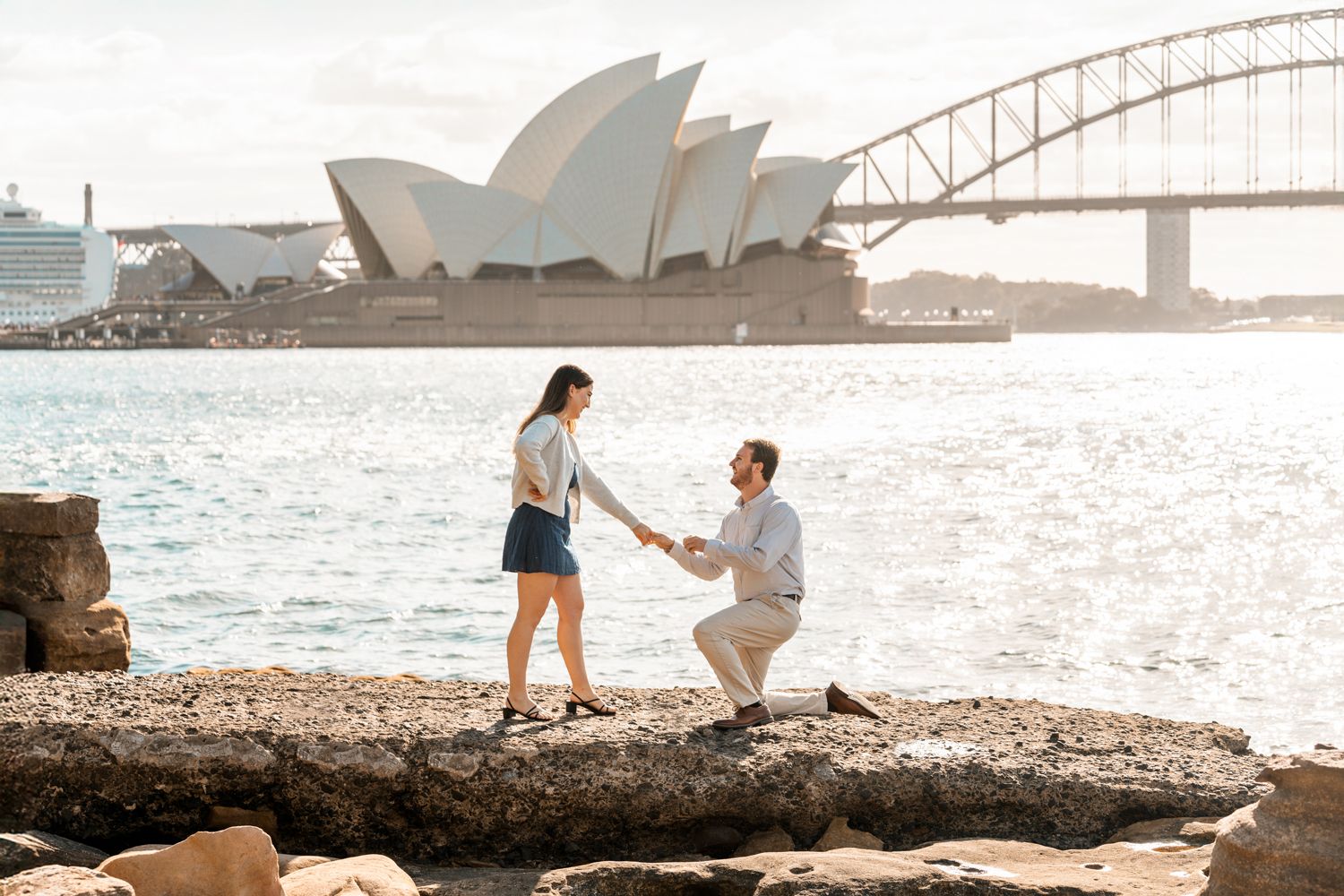 near the water man in a light coloured cloths is kneeling in front of a women holding her hand to propose her, during a secret proposal
