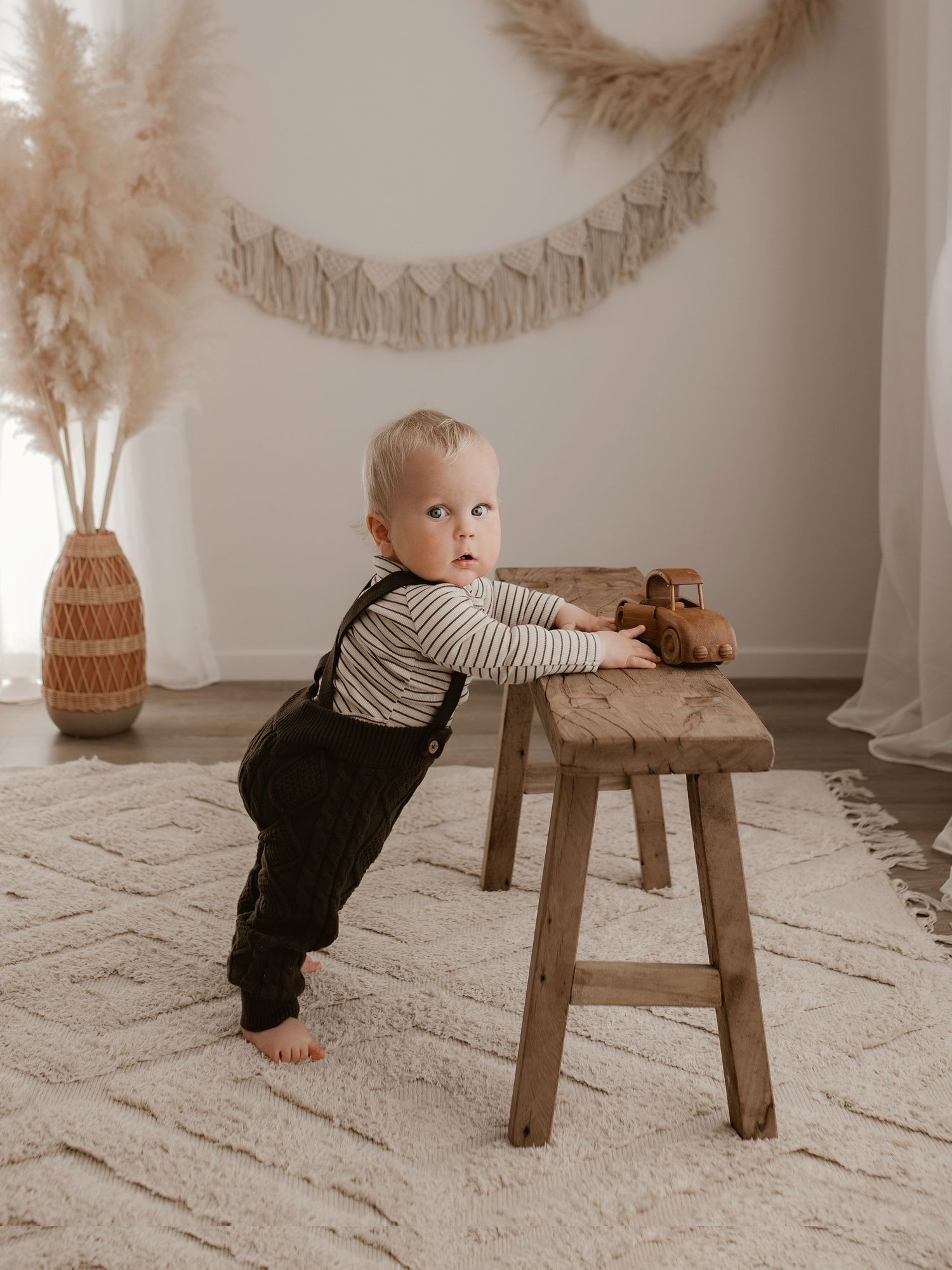 A small child in striped shirt and overalls learns to stand while leaning on a wooden stool in a neutral toned nursery.