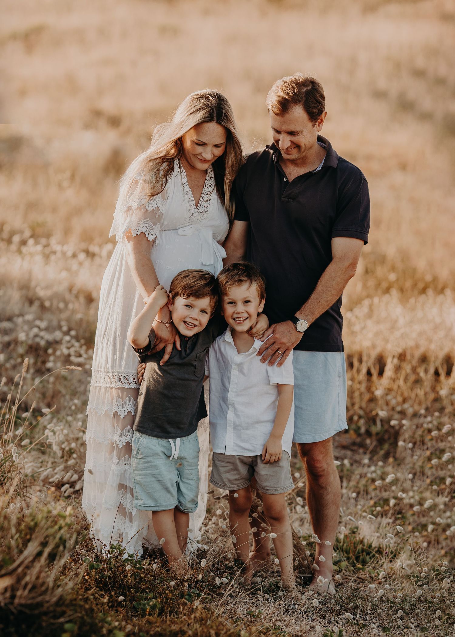 Family of four in coordinating casual outfits posing together in golden field at sunset.