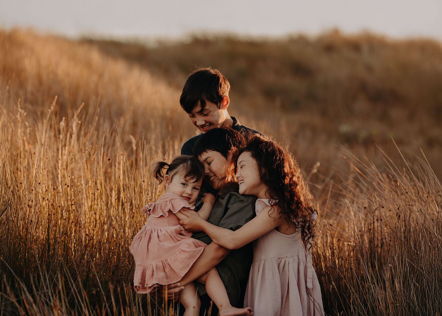 Family of four in coordinating casual outfits posing together in golden field at sunset.