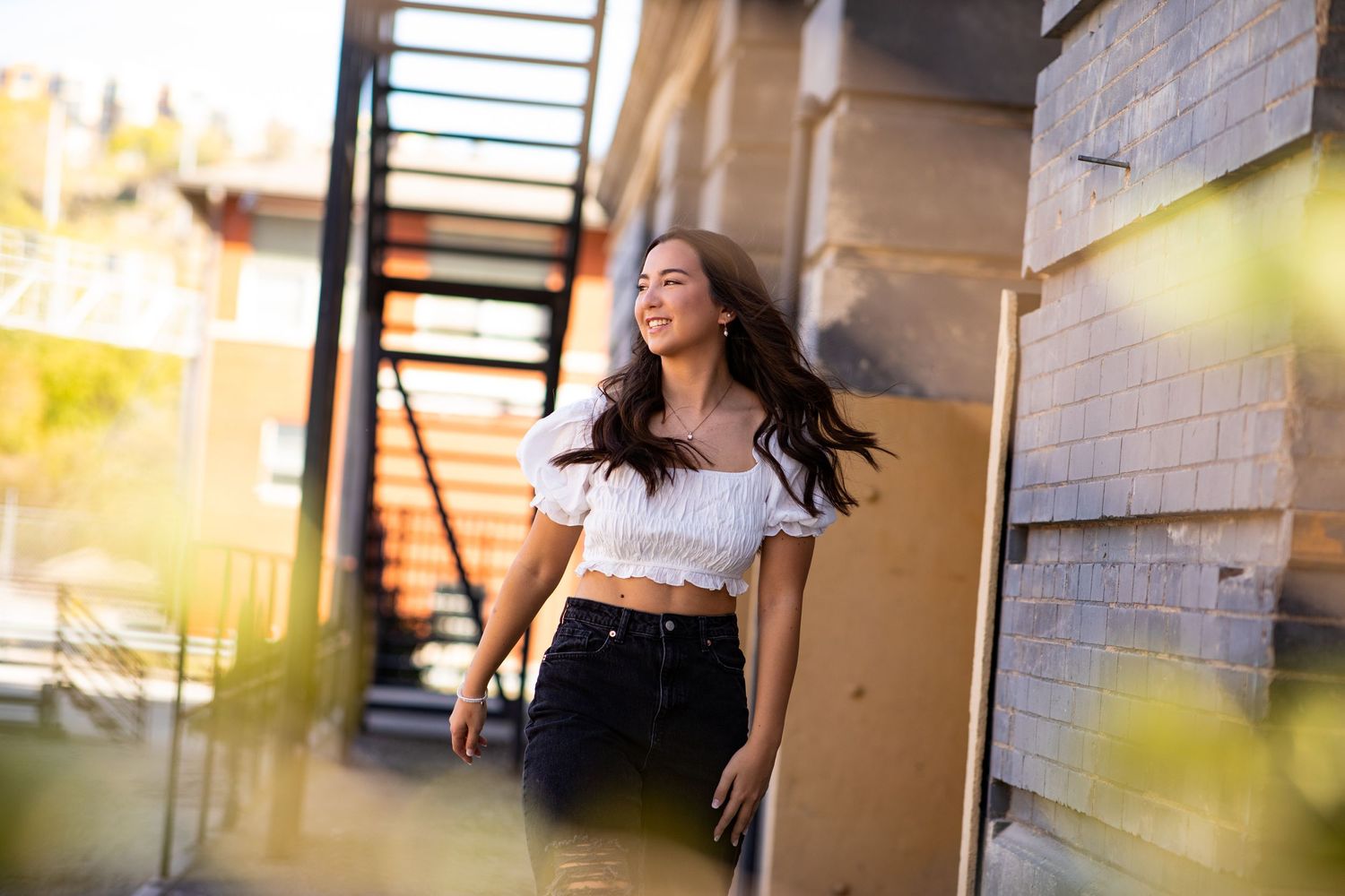 A person in a white crop top and black pants smiles while standing against a sunlit wall with blurred foreground elements.