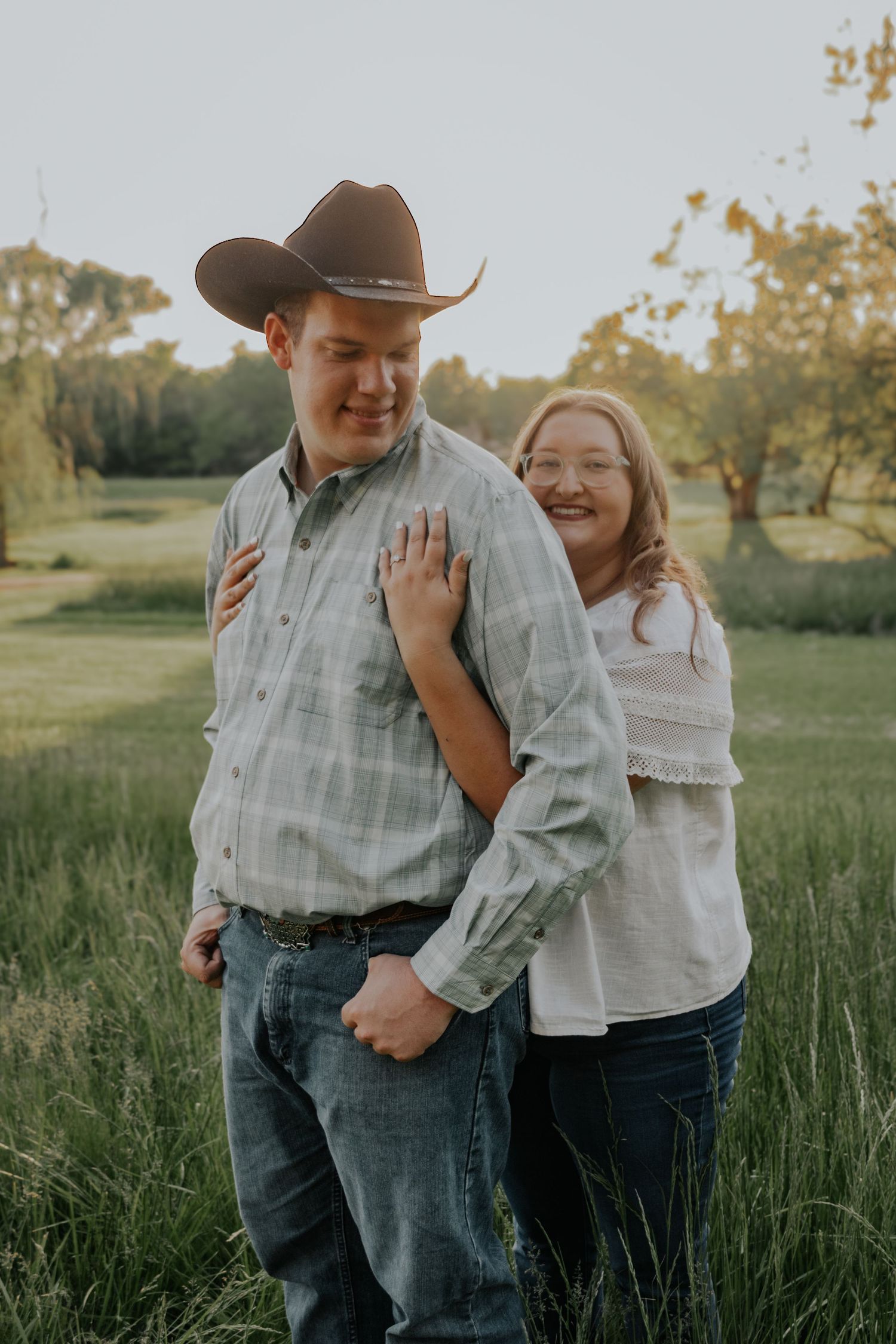 A couple shares a joyful moment together in a grassy field at sunset, with the person wearing a cowboy hat and plaid shirt.