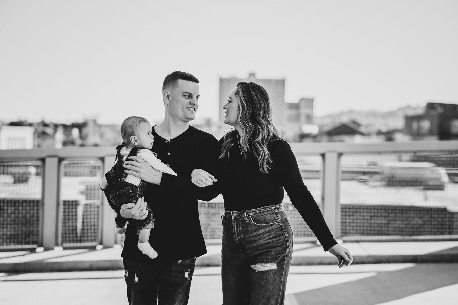 Black and white photo of couple with baby on rooftop terrace with urban backdrop.
