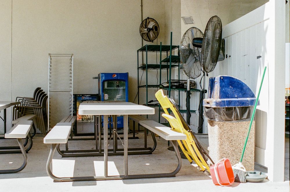 A cluttered garage interior with picnic tables, garden tools, stacked chairs, and storage shelves against white walls.