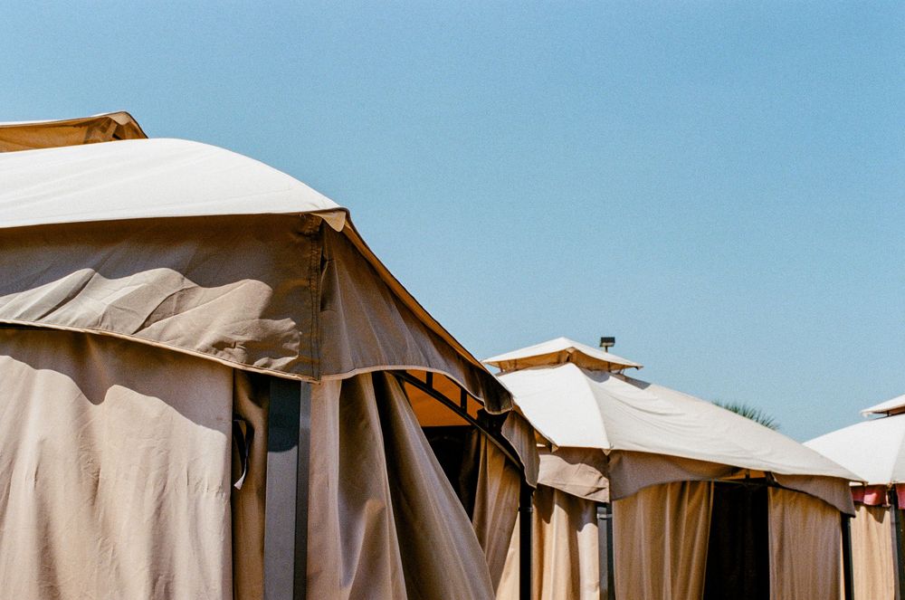 Row of beige canvas glamping tents with white covers against bright blue sky at luxury outdoor resort.