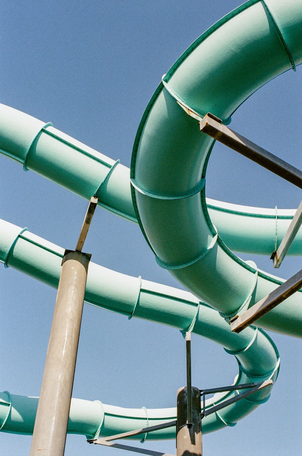 A curved green water slide tube against a blue sky, supported by metal poles at a water park.