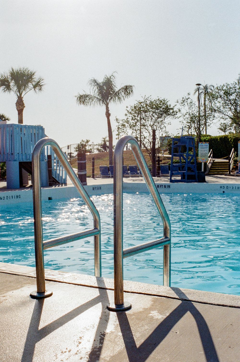 Swimming pool steps and handrails lead into blue water with palm trees and a waterslide visible in the background.