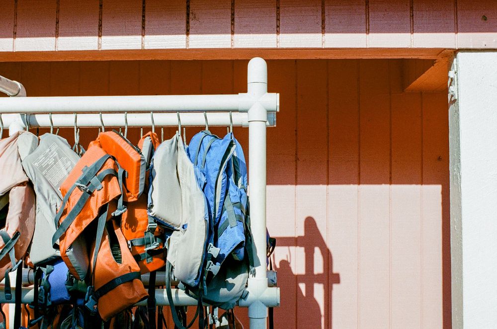 Backpacks and sports gear hang on a white rack against a coral-colored wall in outdoor lighting.