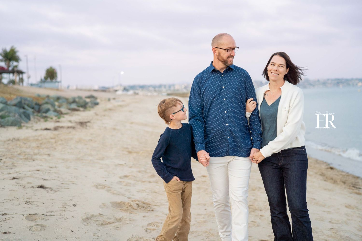 Family walking together on sandy beach with boats visible in harbor background.
