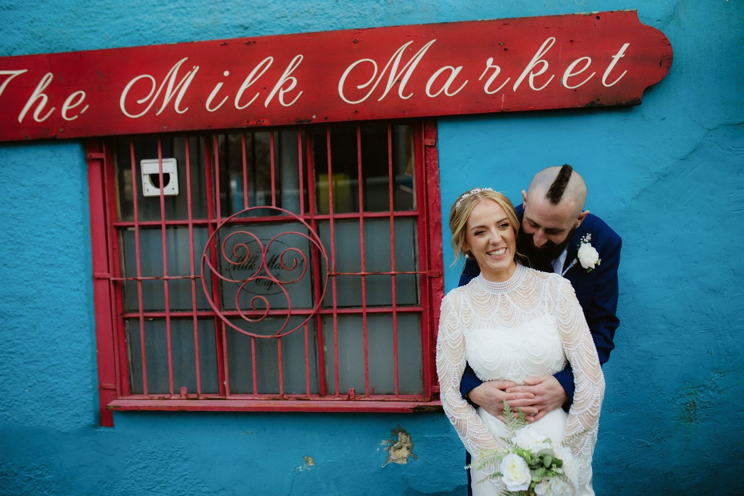 A couple poses in front of The Milk Market sign with vibrant turquoise walls and red trim in an intimate moment.