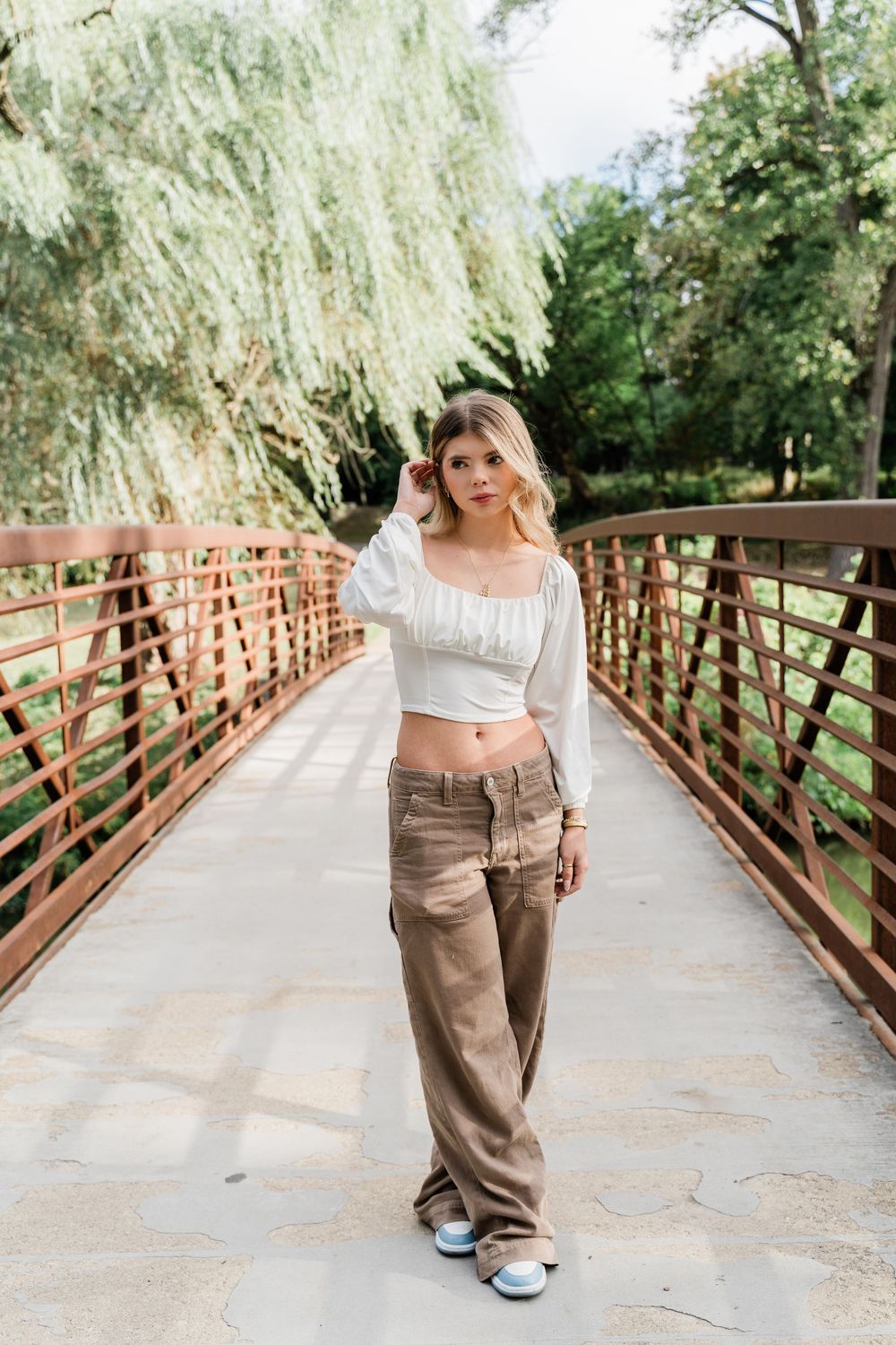 A person poses on a wooden bridge wearing a white crop top, khaki cargo pants and sneakers beneath a willow tree.