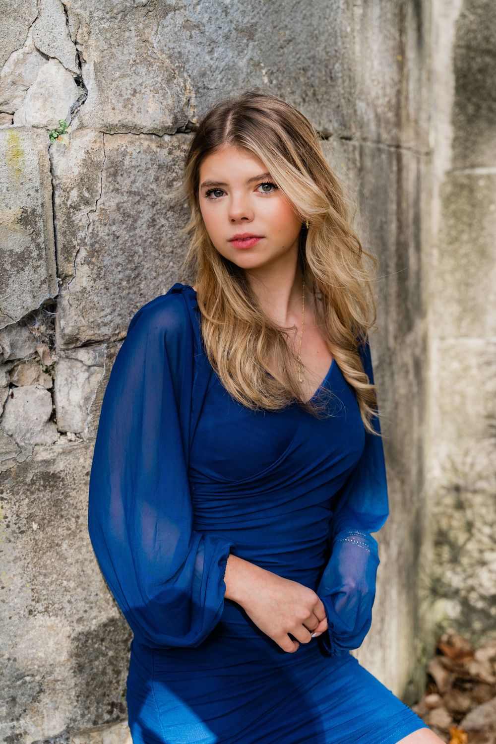 A person in an elegant royal blue dress with sheer sleeves poses against a weathered stone wall.