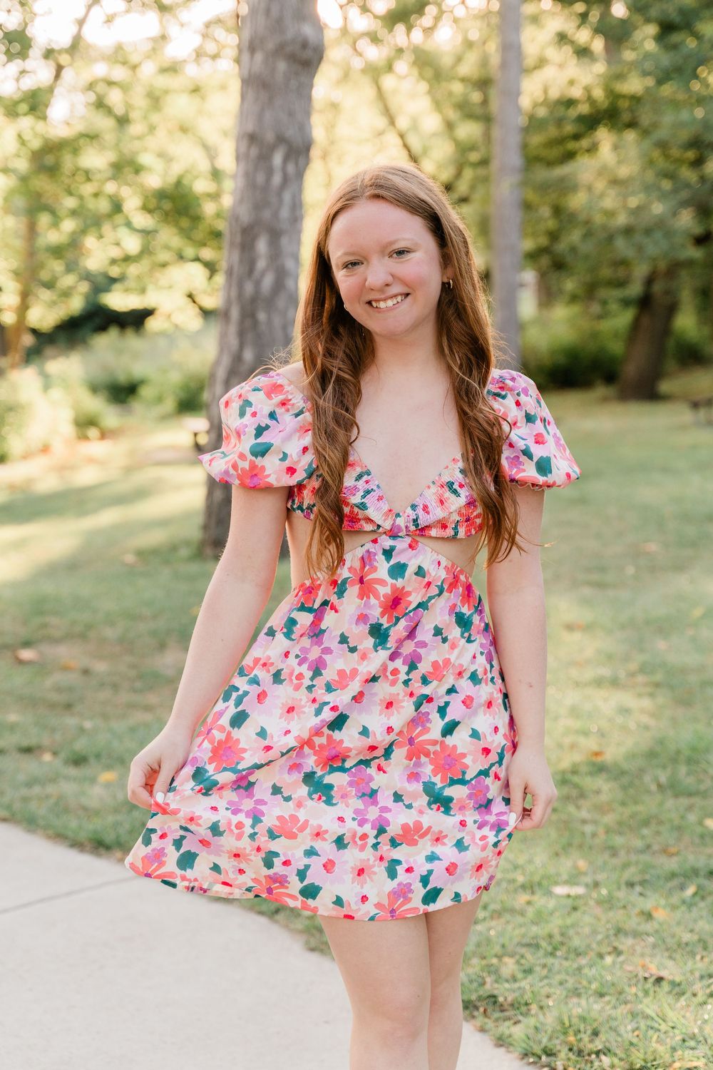 Someone wearing a pink floral print dress with puff sleeves poses on a walkway surrounded by trees in summer.