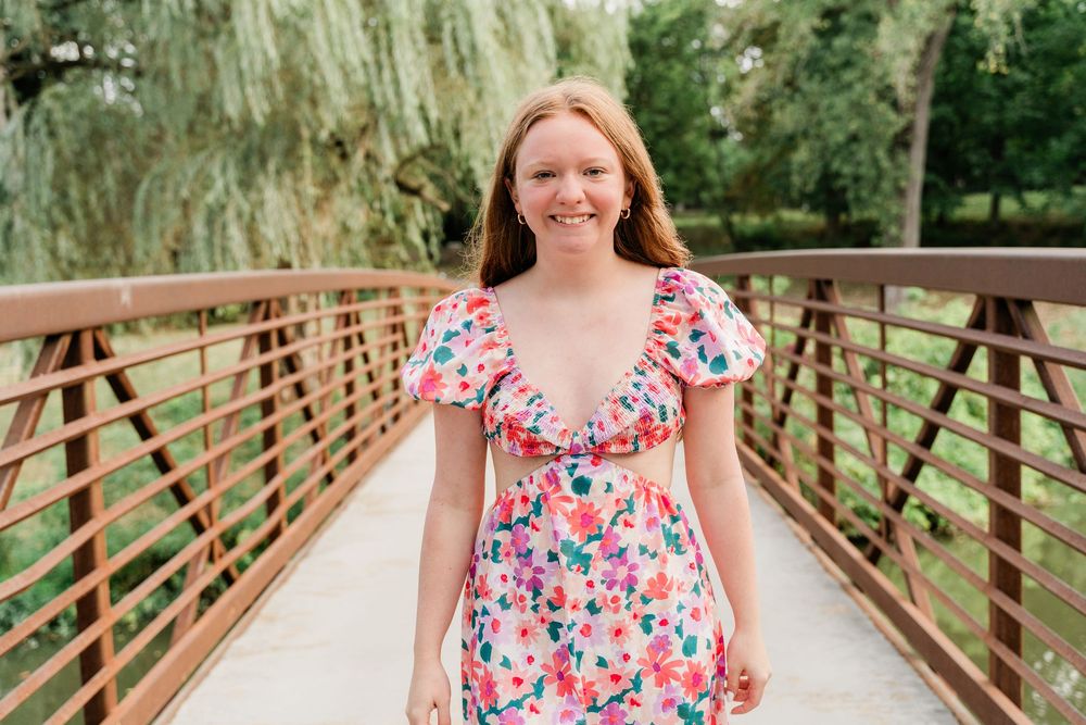 A floral summer dress with ruffled neckline on a wooden bridge surrounded by weeping willow trees in the background.