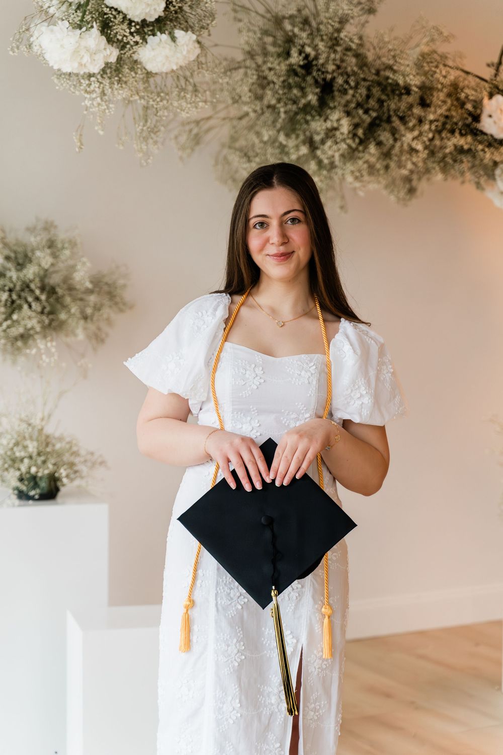 Graduate in white dress holds black graduation cap under floral decor in elegant indoor setting.