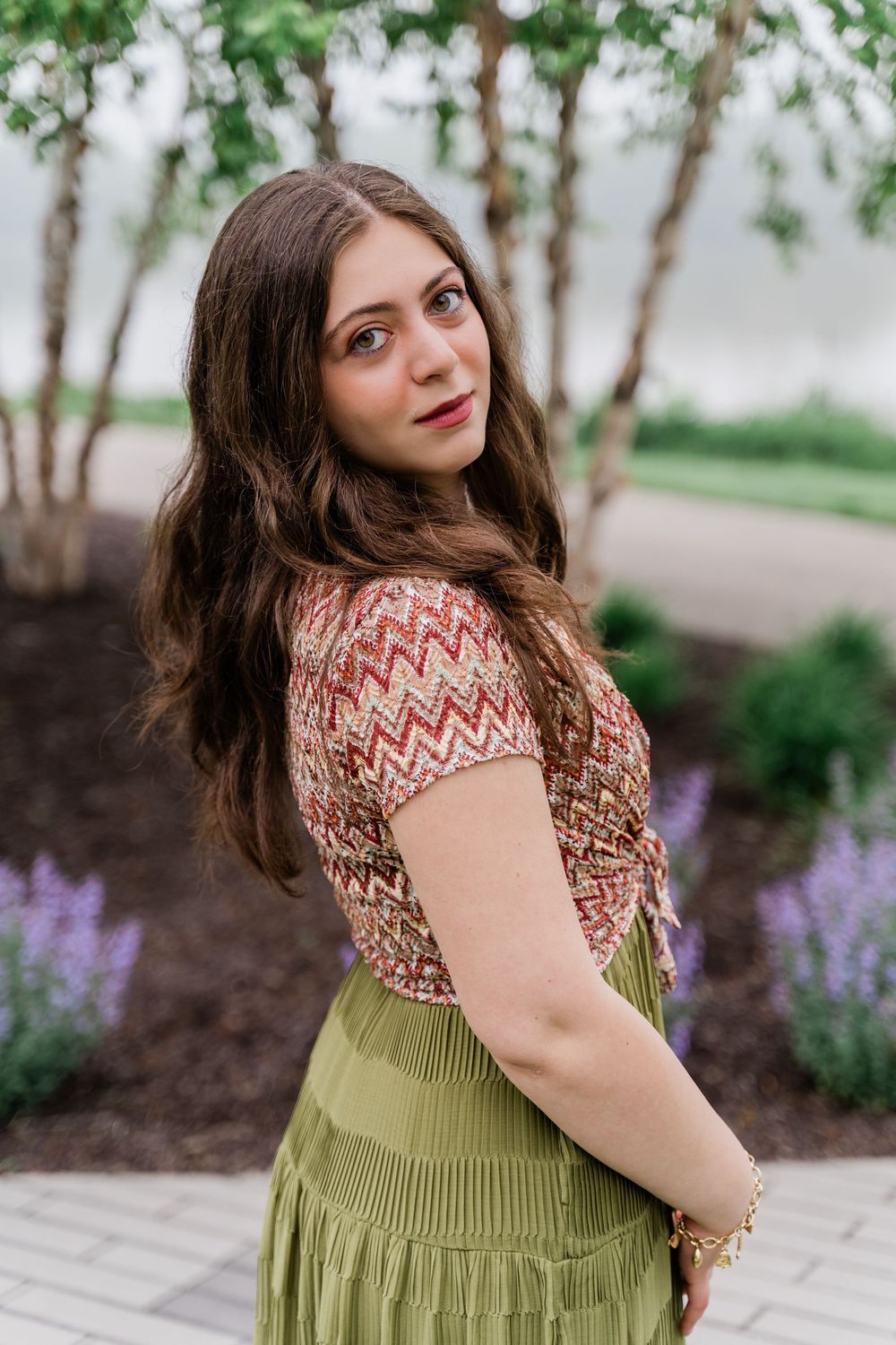 Woman in patterned top and olive pleated skirt poses outdoors near blooming purple flowers and trees.