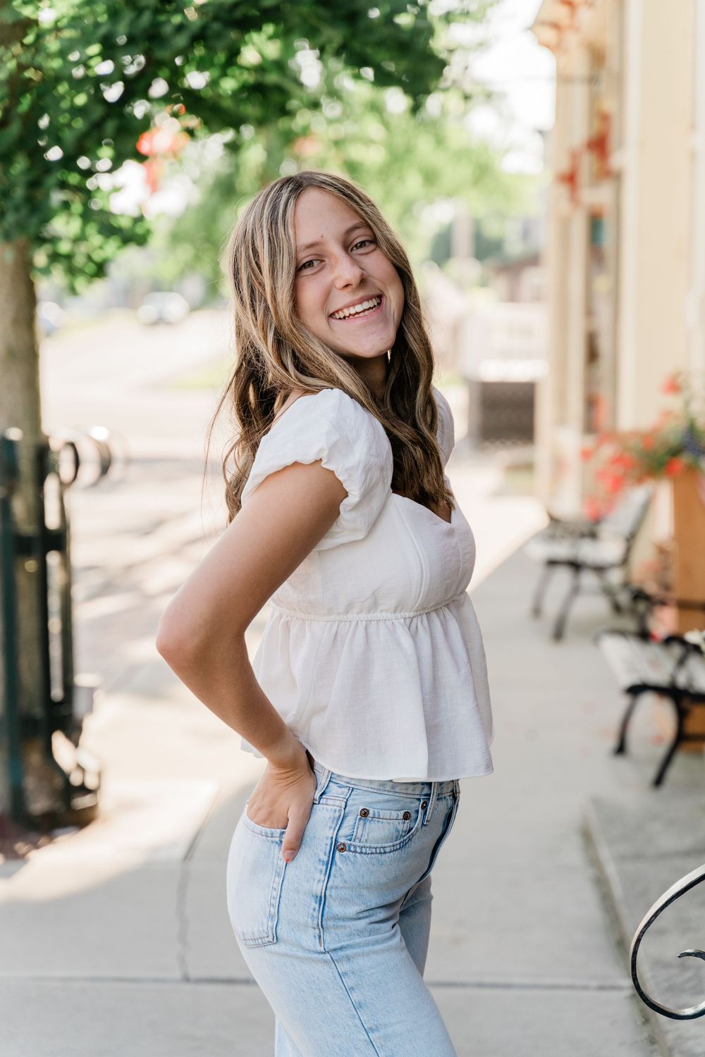 Person in white peplum top and light jeans poses cheerfully on a sidewalk with trees and storefronts in background.