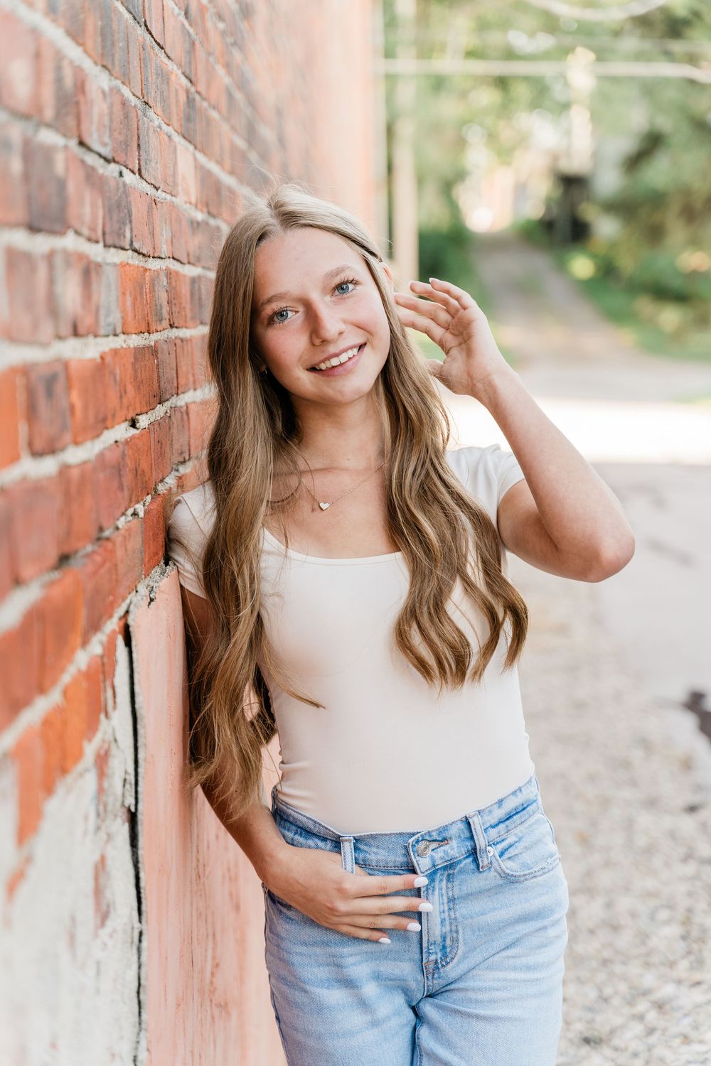 Casual portrait against brick wall featuring light brown wavy hair, white top and light denim jeans outdoors.