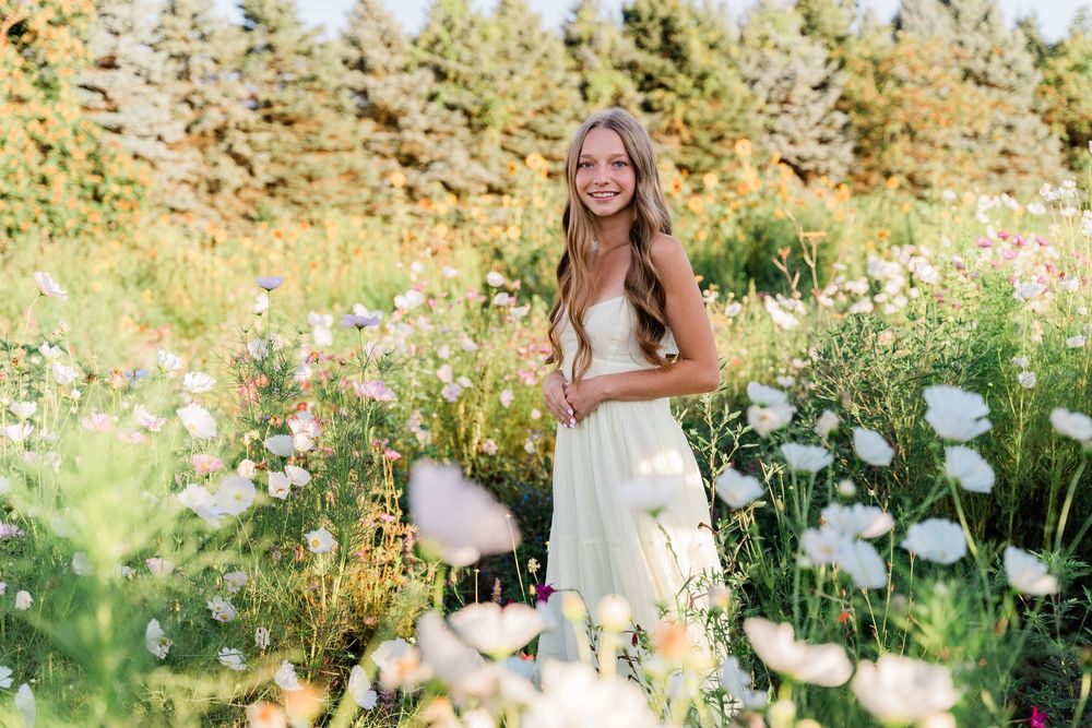 Person in a white dress stands surrounded by blooming white flowers and greenery in a sunny garden setting.