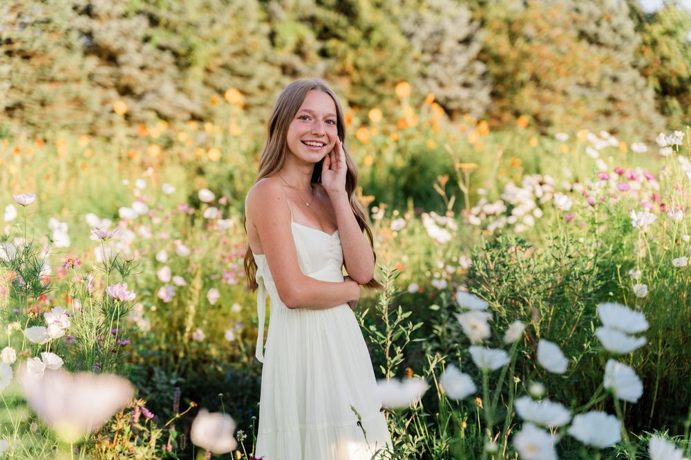 A woman in a flowing white dress poses joyfully in a wildflower meadow with white blooms on a sunny summer day.