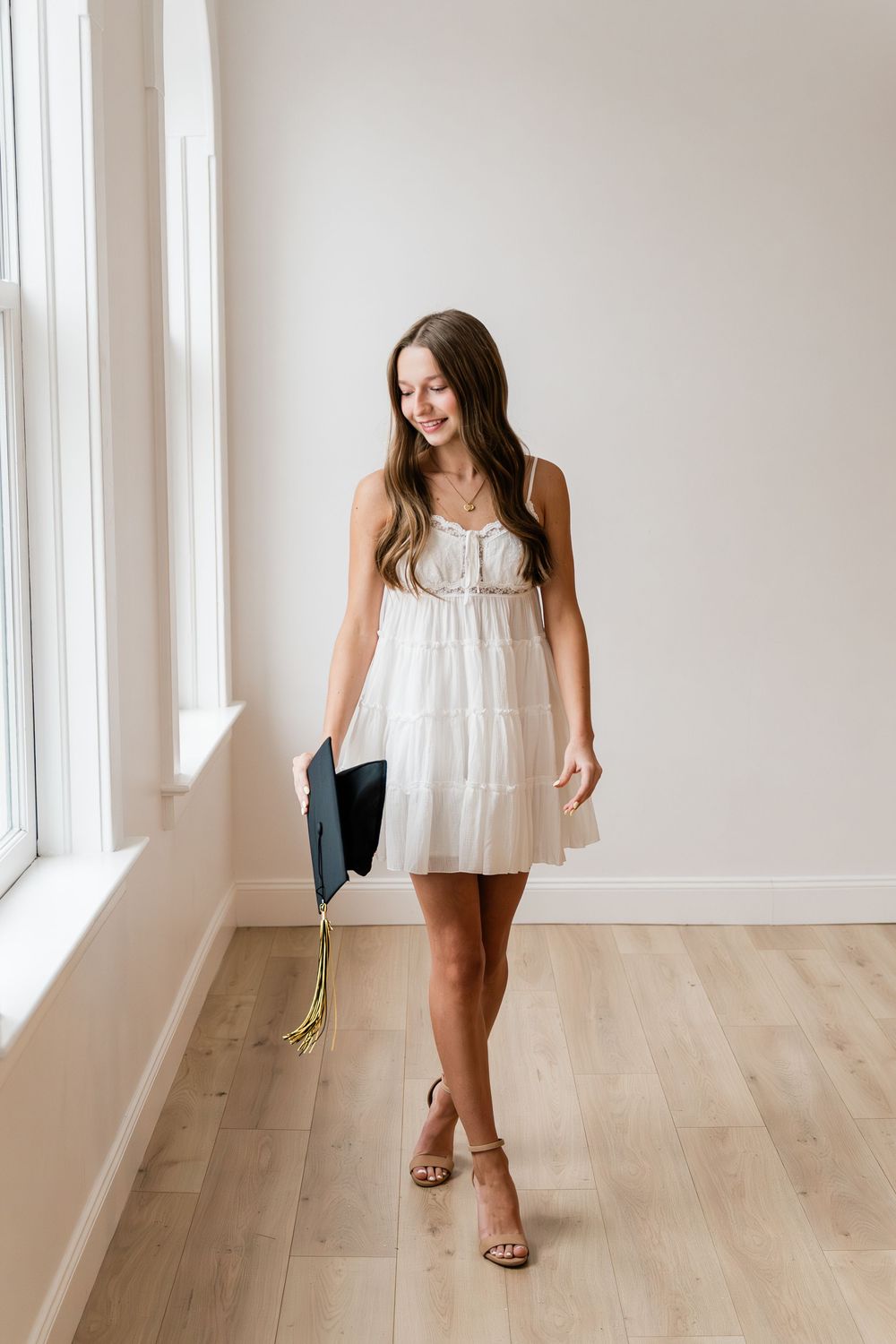 Young woman in flowy white sundress and sandals poses by window in bright minimal interior with hardwood floors.