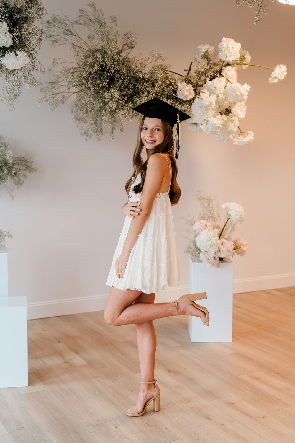 A graduation portrait with white dress and cap against floral decorations and light wood flooring.