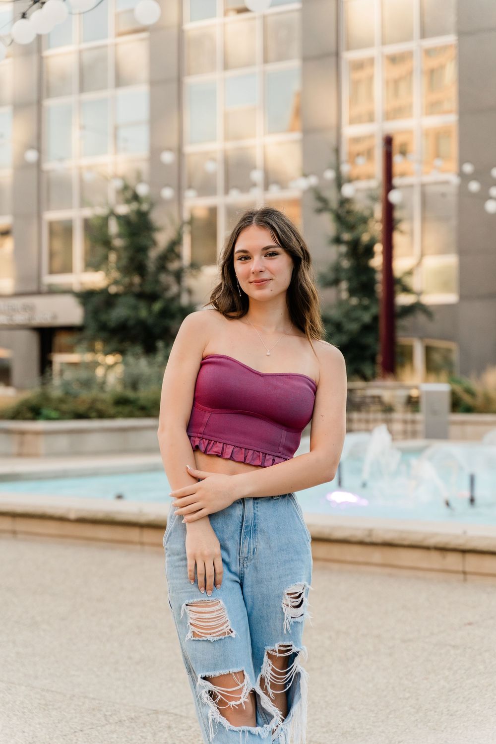 A woman in a purple tube top and distressed jeans poses near a fountain outside a modern building.