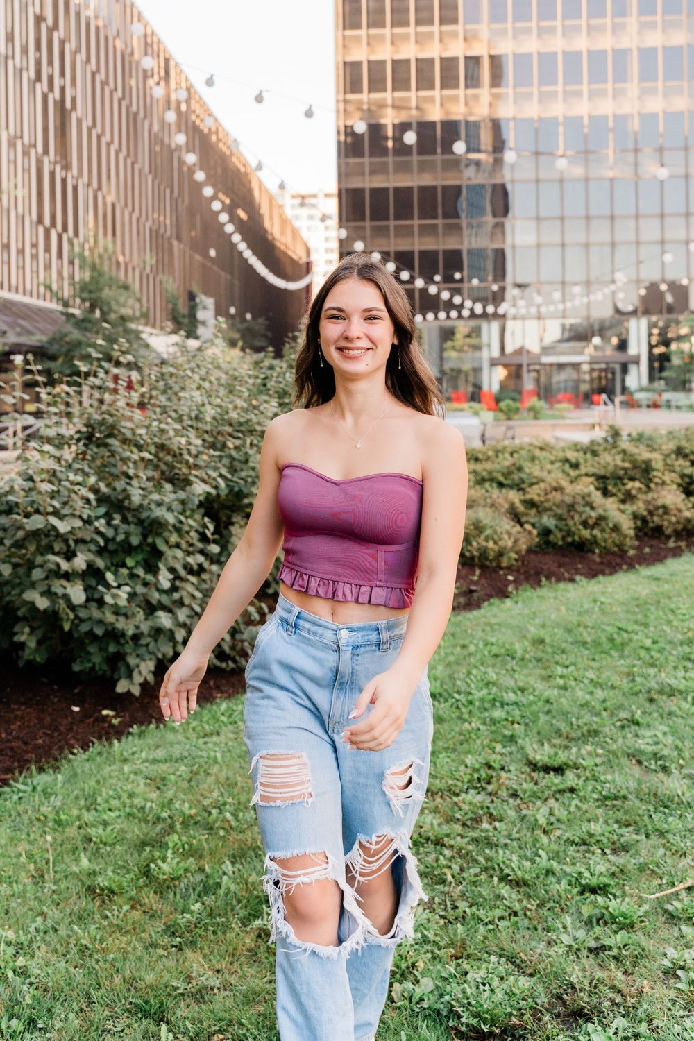 Young woman in purple crop top and ripped jeans standing in front of urban garden and glass building.