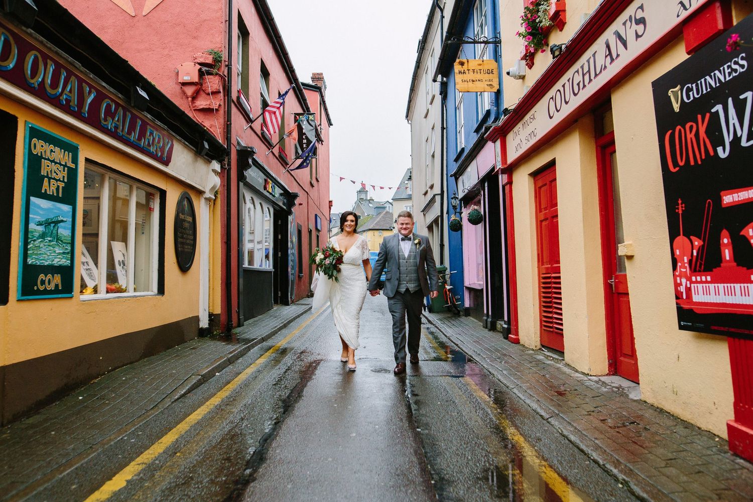 Two people walk down a narrow rainy street in Kinsale, Cork lined with colorful storefronts in a charming Irish town.