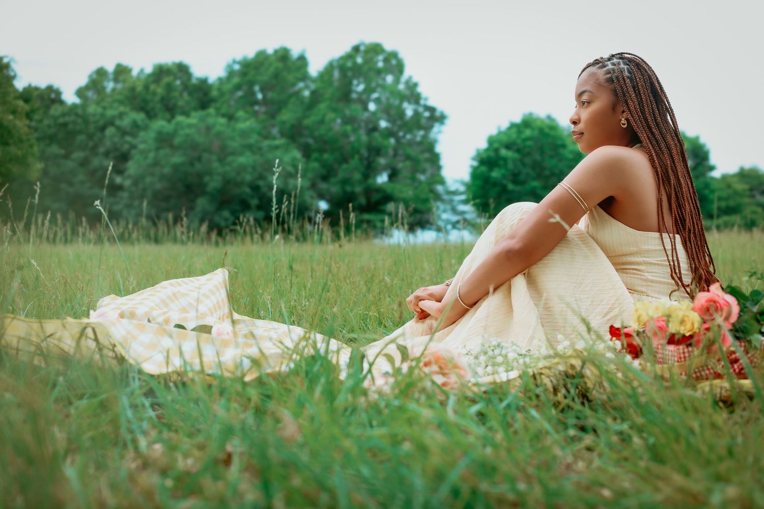 Young Black woman in a white strapless dress sits in a grassy meadow with braided hair and flowers.