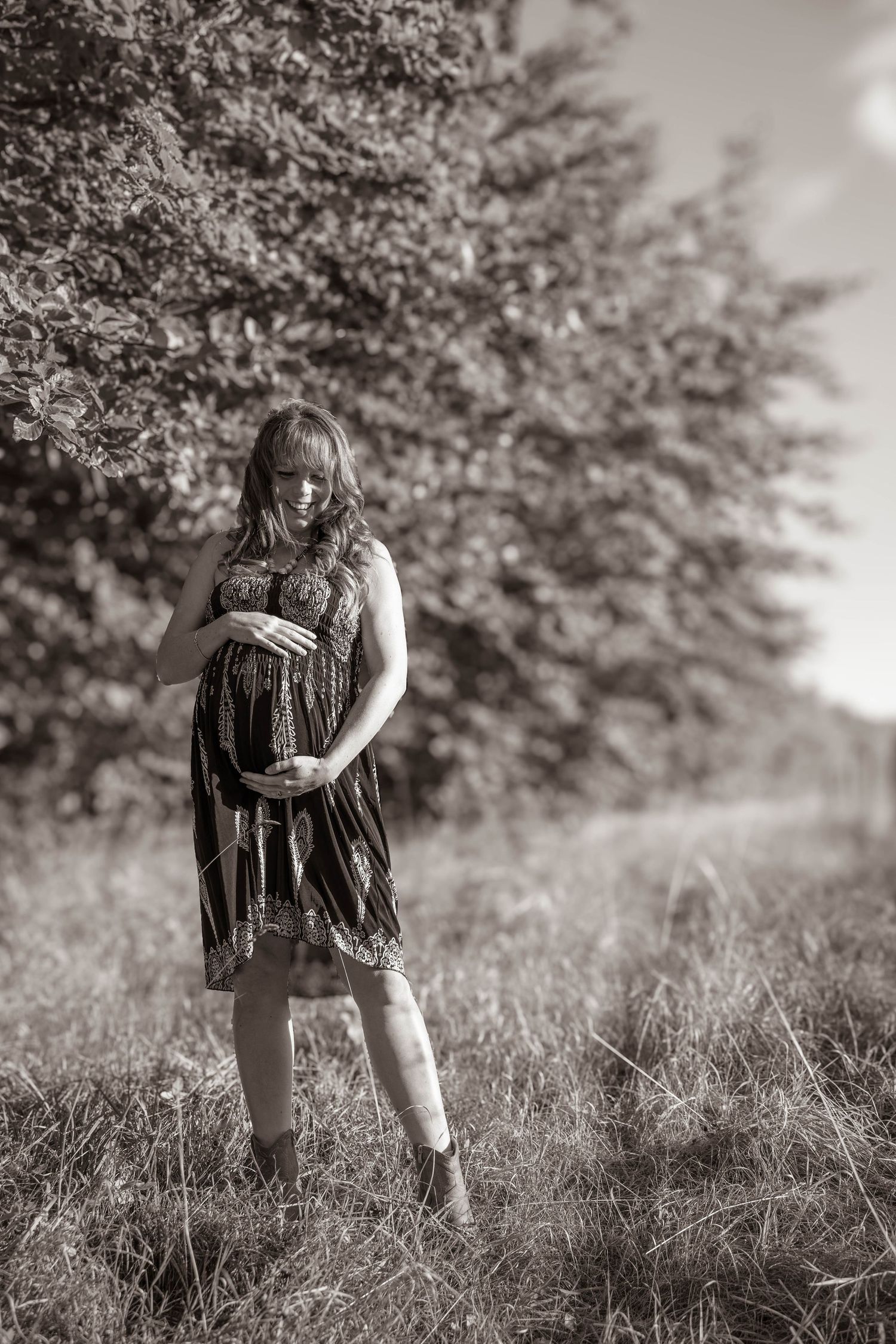 A person in a flowy dress stands in a sunlit meadow during a maternity photoshoot.