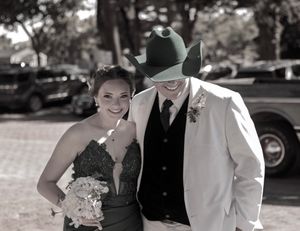A formal couple in prom attire pose together outdoors, with the man wearing a light colored suit and green hat.