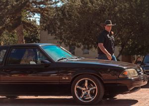 Black Ford Mustang Fox Body with chrome wheels parked on dirt road under trees in warm sunlight.