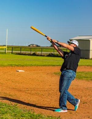 Baseball player in jeans and black jacket swings a yellow bat on a dirt field with green grass and blue sky background.