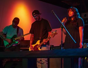 Musicians perform on stage under dramatic orange and blue lighting with guitar players and a vocalist at microphone stand.