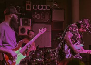 Musicians perform on stage in a dimly lit room with bass and guitar players silhouetted against purple lighting.