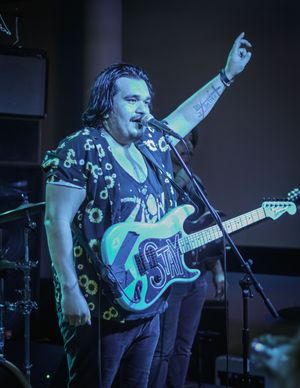 Musician in daisy print shirt performs on stage with a blue electric guitar under dramatic lighting.