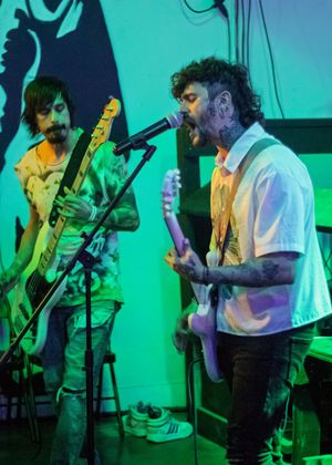 Musicians perform on stage with electric guitars under blue and green lighting during a live concert.