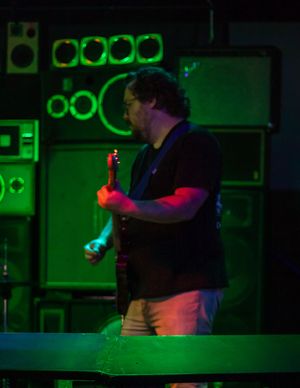 A musician performs on stage illuminated by dramatic green and red lighting with stacks of amplifiers in the background.
