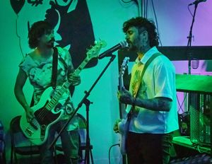 Musicians perform on stage with electric guitars under vibrant green lighting at a concert venue.