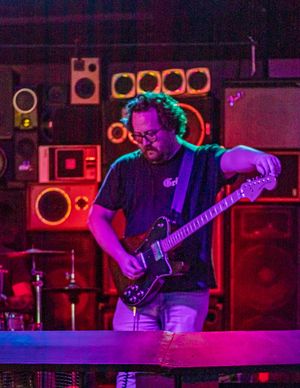 A musician performs on stage with a bass guitar under dramatic purple and blue lighting with speakers in the background.