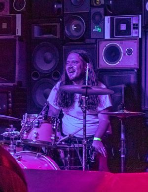 Energetic drummer performs behind a drum kit with a wall of speakers in purple-lit concert environment.