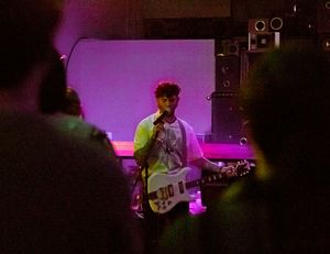 Performer on stage with electric guitar bathed in purple lighting during live concert performance.