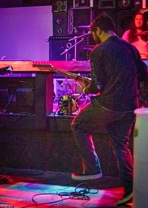 Musician performing on stage with a guitar under purple and pink lighting effects at a live music venue.