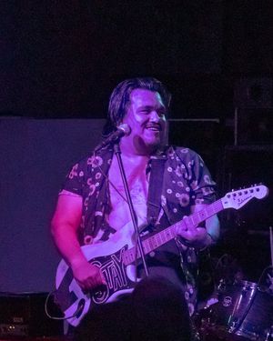 Musician performing on stage with an electric guitar in purple lighting while wearing a floral print shirt.